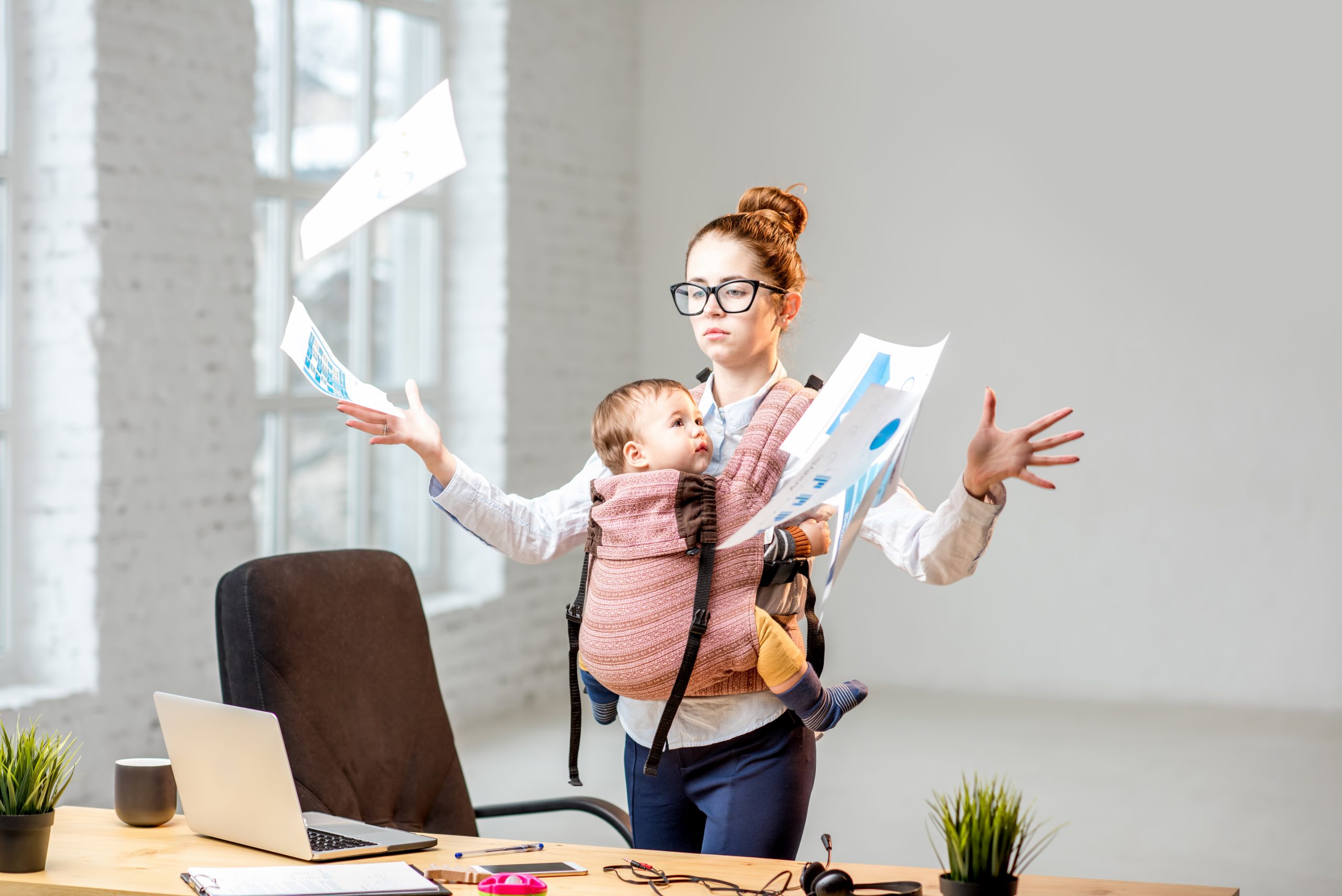 Multitasking and exhausted businesswoman throwing up a documents standing with her baby son during the work at the office